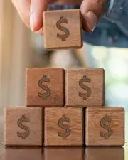 A person stacking wooden blocks that have dollar signs on them.